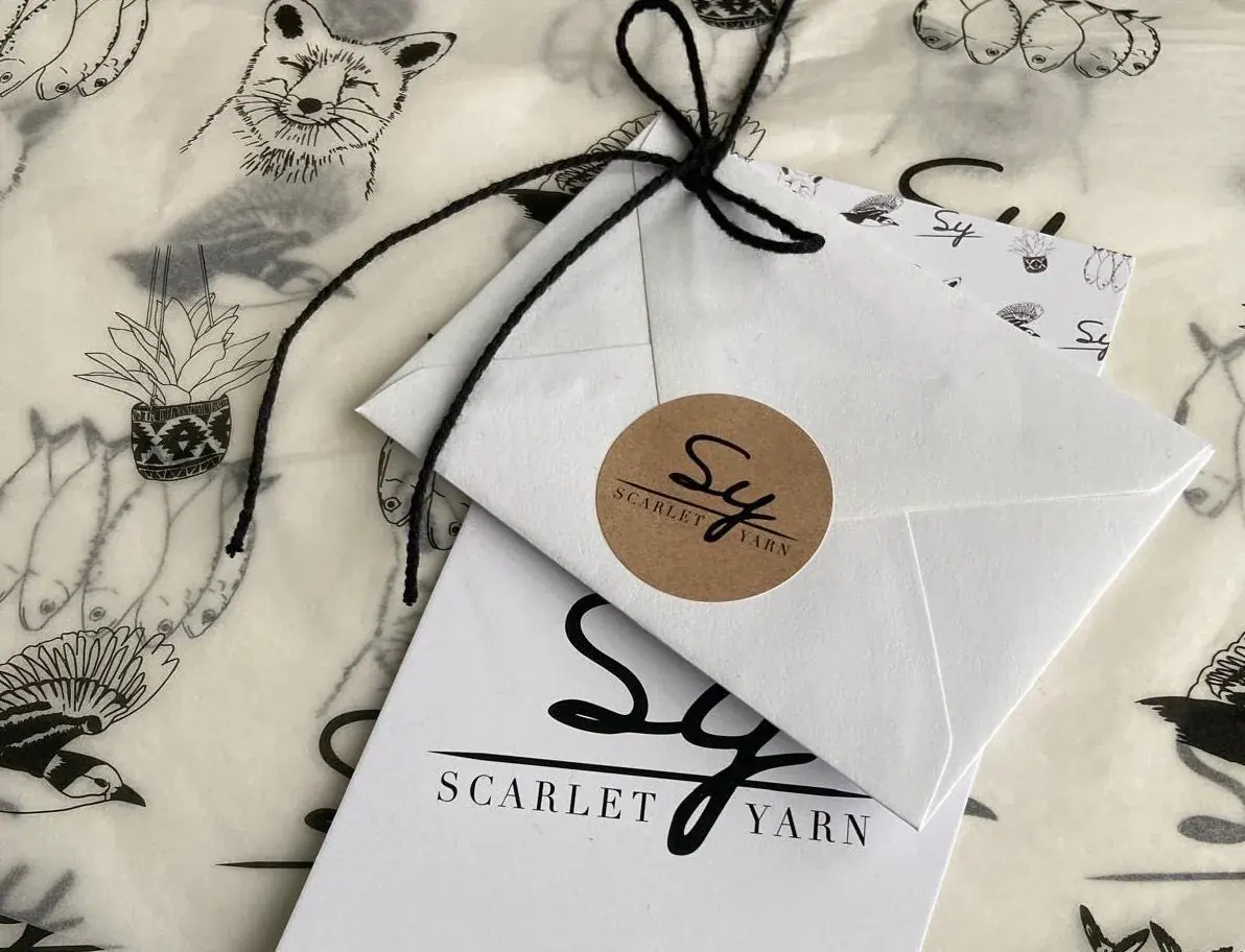 A Scarlet Yarn gift: a signed card and envelope sealed with a kraft Sy sticker, tied with black twine on tissue printed with Rachael's drawings