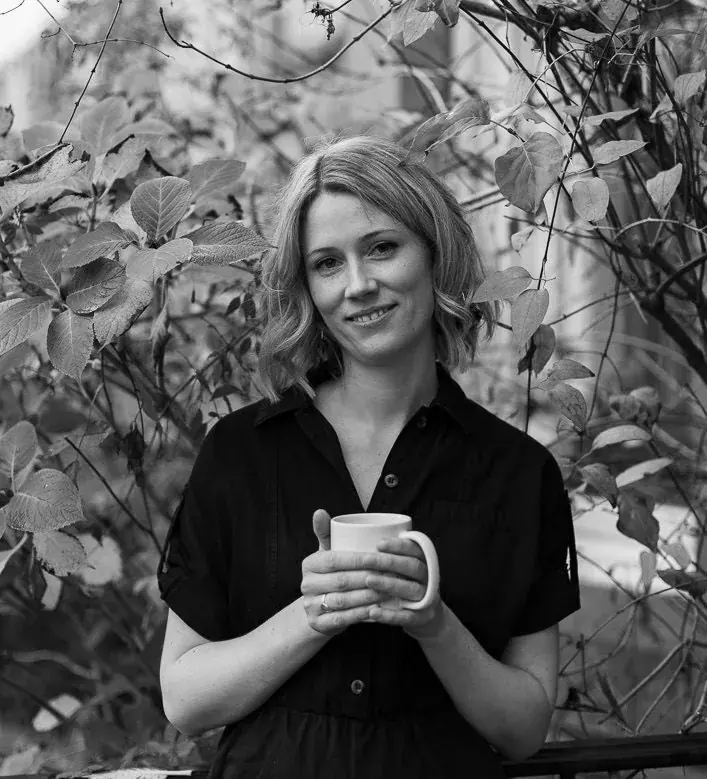 Rachael, the designer behind Scarlet Yarn, holding a mug in front of leafy plants